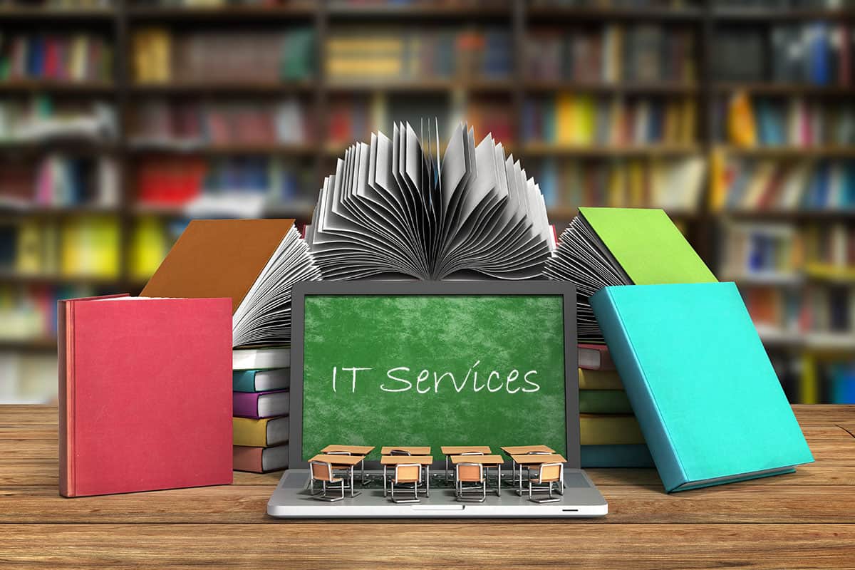 A laptop sits on a school desk, surrounded by books. The screen displays a chalkboard with the text "IT Services" written on it.