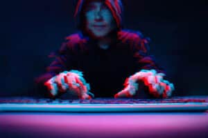 A person in a dark hoodie is typing on a keyboard, with a blurred effect giving a sense of movement. The scene is illuminated with blue and red tones, enhancing the mysterious atmosphere.