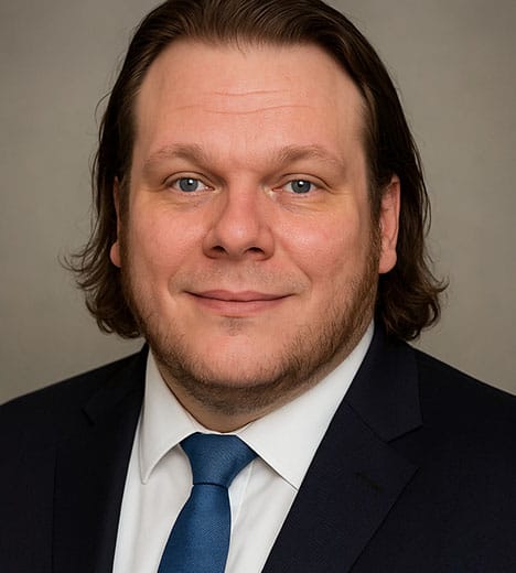 A professional headshot of a man with shoulder-length dark hair, wearing a black suit, white shirt, and a blue tie, smiling softly against a neutral background.