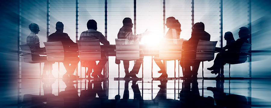 Silhouettes of people sitting around a conference table, engaged in discussion, with light streaming through blinds in the background.