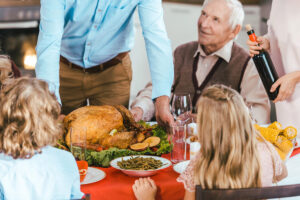 A festive dining table with a roasted turkey at the center, surrounded by various side dishes. A man in a blue shirt serves the turkey while an elderly man in a sweater looks on. Children with curly hair and a girl with long hair sit at the table, watching the scene. A woman pours wine from a bottle.