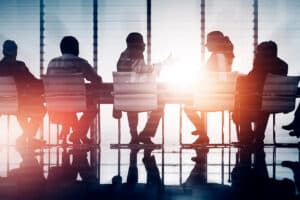 Silhouettes of six individuals seated around a conference table, with sunlight streaming through large windows, creating a dramatic effect on the scene.