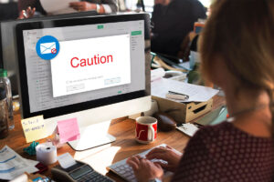 A person is typing on a keyboard in front of a computer displaying an email warning message that says "Caution" in red. The desk is cluttered with paperwork, a red mug, and various office supplies.