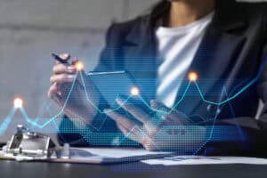 A person in a black blazer holds a smartphone in one hand and a pen in the other, surrounded by digital graphs and data visualizations on a blurred background. Papers and a clipboard are visible on the table.