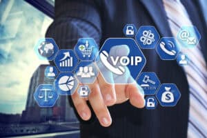 A businessman in a suit is reaching out to touch a virtual interface displaying various icons, including a prominent "VOIP" symbol, along with graphics representing communication, finance, and data analytics. The background features a modern office setting.