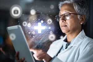 A healthcare professional in a lab coat examines a tablet, with digital health icons and a glowing medical cross graphic overlay.